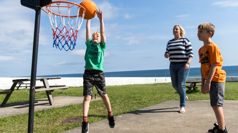 A family in summer clothes plays basketball in the sunshine within the whitewashed stone walls of the Foghorn Field. A child is mid-jump as they throw the orange ball into the net.